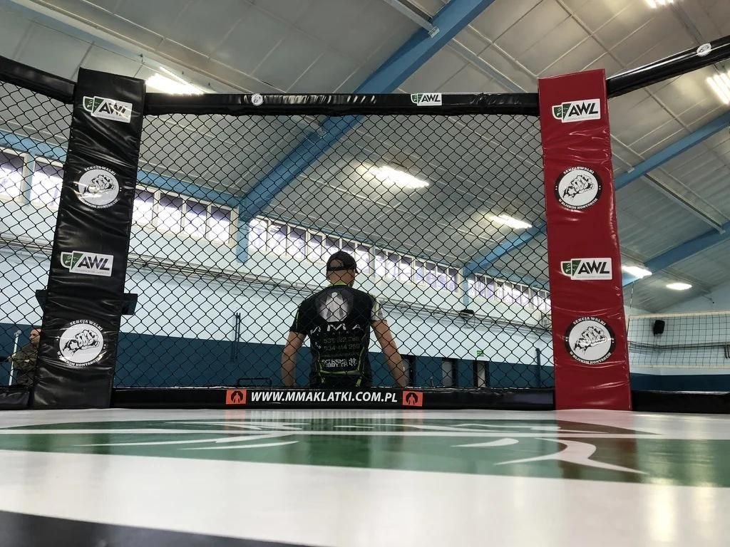 Klatka MMA CLASSIC w hali sportowej z bliska: czarny i czerwony słup z logotypami sponsorów, siatka i biała mata z nadrukiem, trener w środku