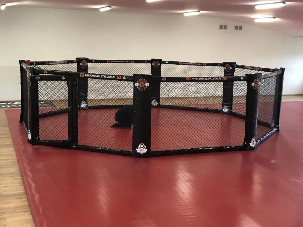 Ośmiokątna klatka MMA CLASSIC na poziomie posadzki, czarne obicia słupów i bandy z logotypami, czerwona mata w sali treningowej