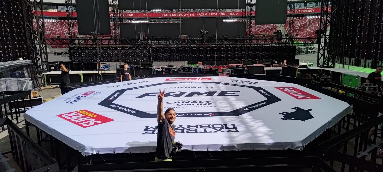 Gotowy oktagonalny podest klatki MMA przykryty brandowanym pokrowcem FAME MMA, w tle trybuny PGE Narodowego.