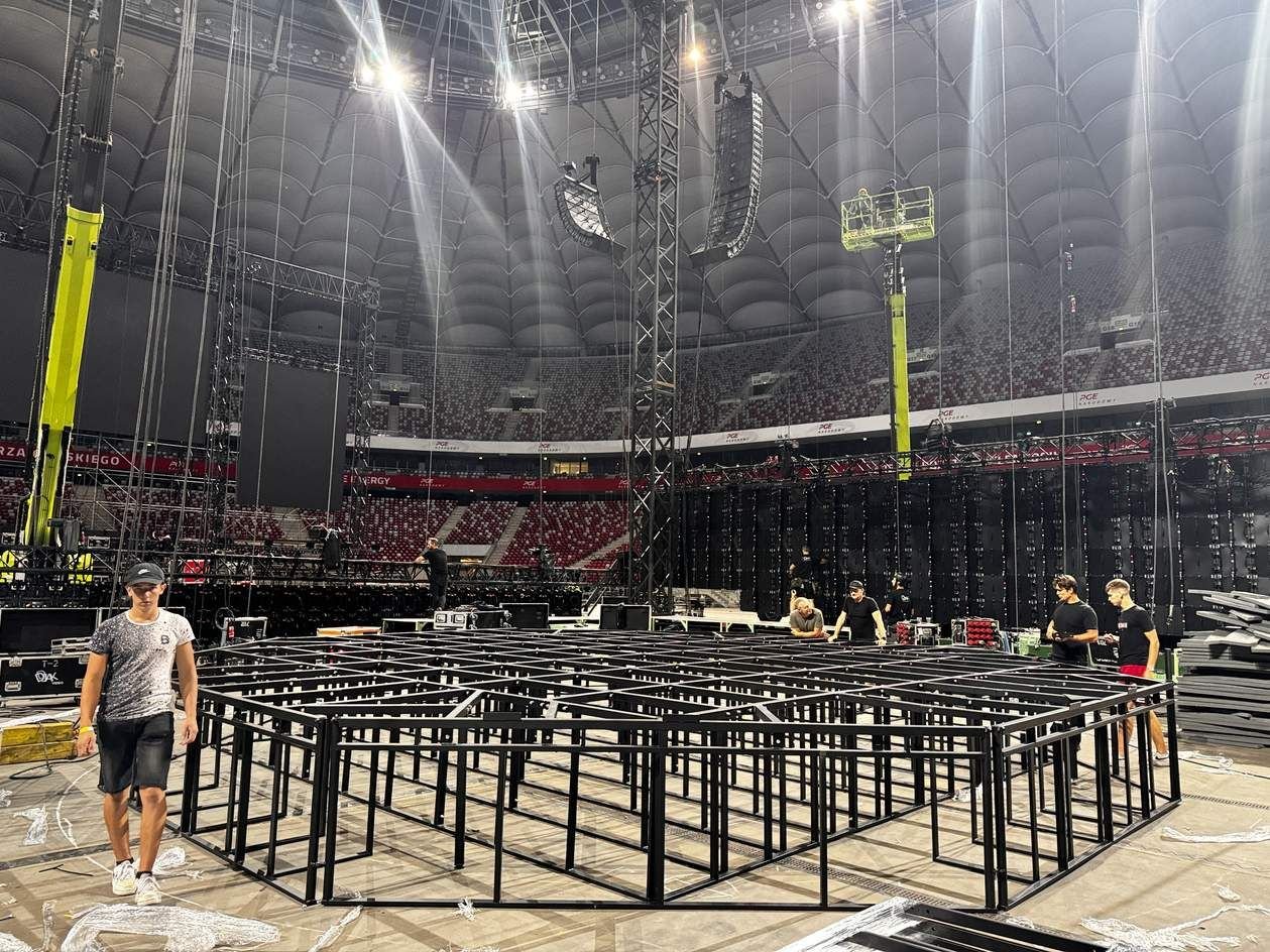 Etap budowy: stalowa konstrukcja nośna podestu klatki MMA na płycie stadionu, ekipa montażowa przy pracy.