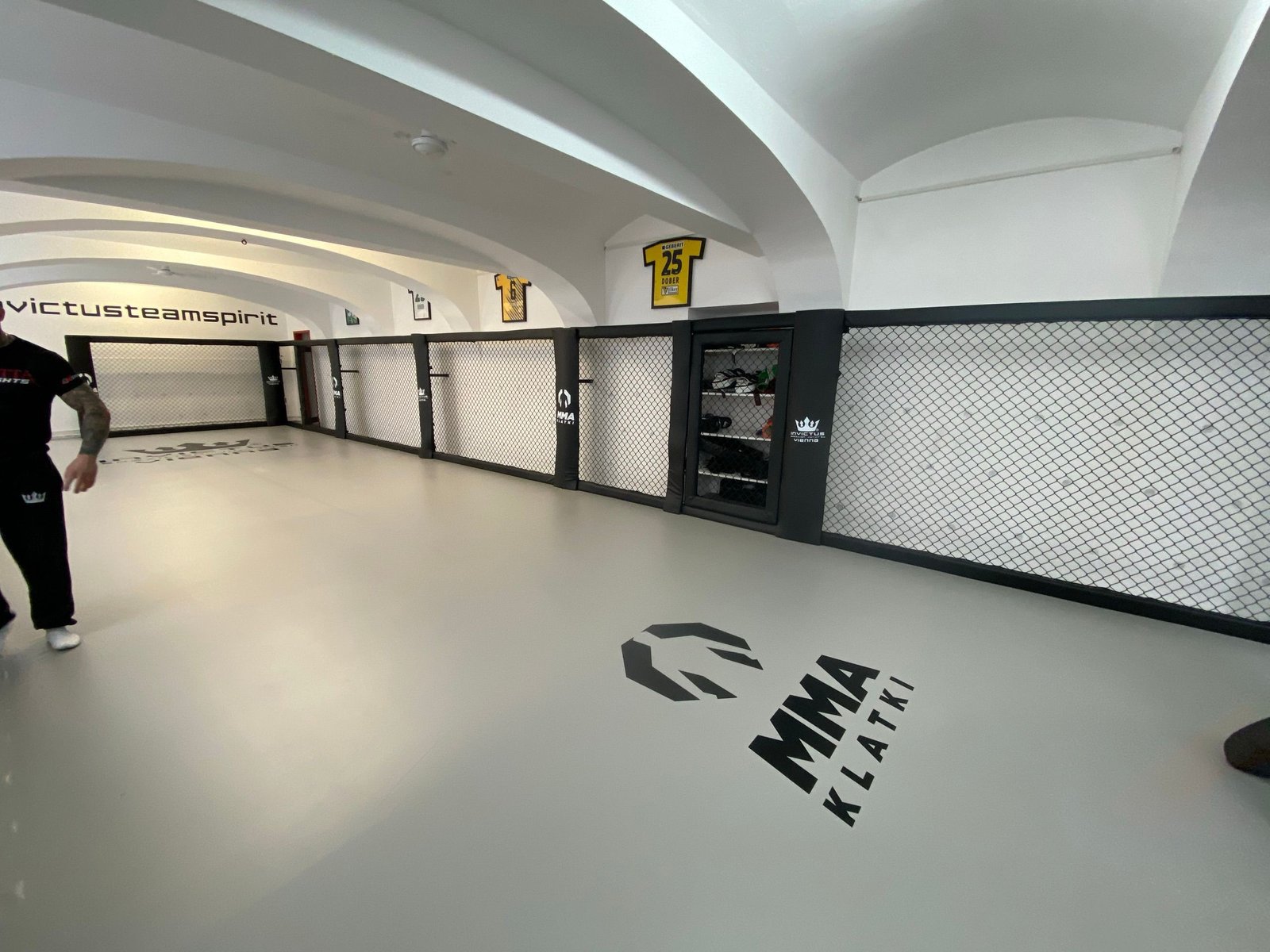 Długa sala treningowa Invictus BJJ Vienna z szarą matą z nadrukami logotypów i ścianą w systemie Cage Wall