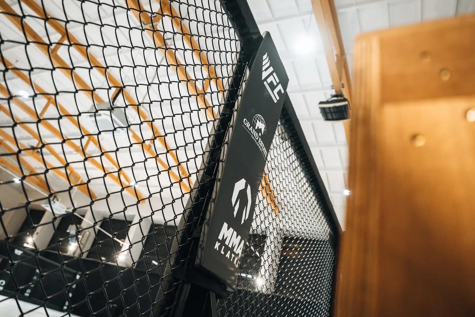 Zbliżenie osłony słupka Cage Wall z logo klubu i sponsora, czarna siatka, drewniane dźwigary hali w tle