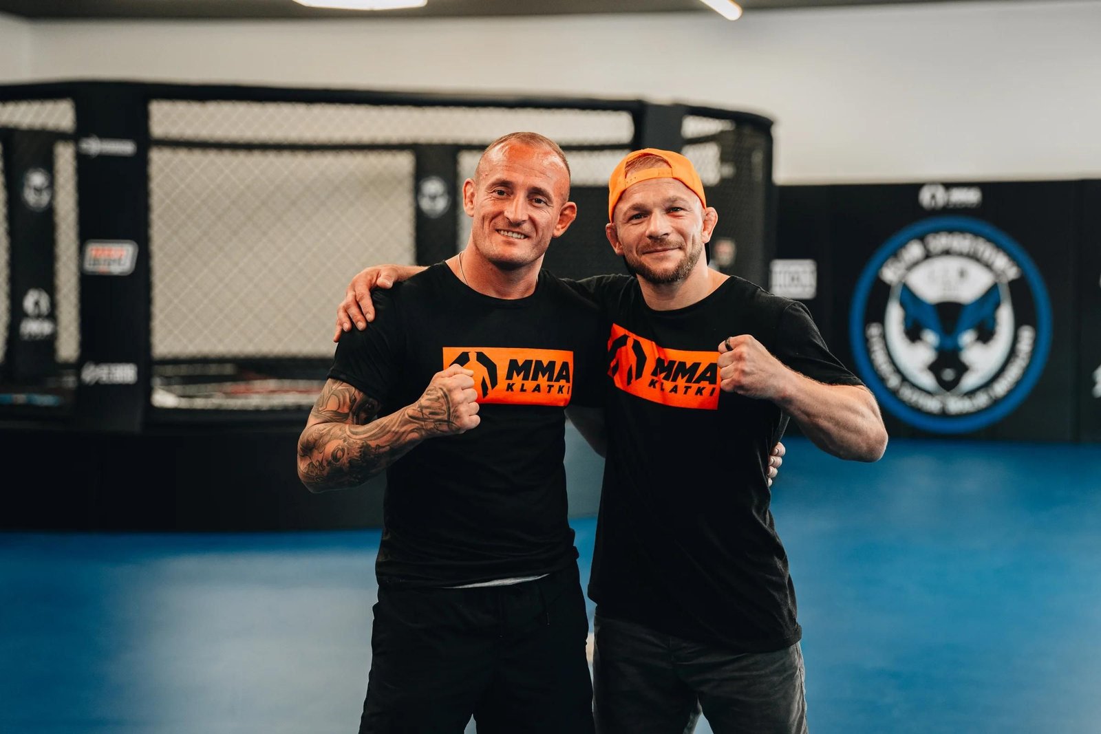 Właściciel klubu i monter MMA Klatki na niebieskiej macie, w tle klatka MMA i czarne maty ścienne z logo Strefy Sztuk Walki Mogilno