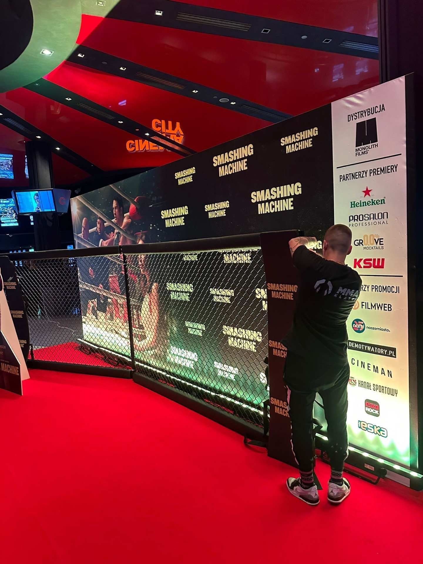 Ujęcie pionowe z foyer kina Cinema City. Na czerwonym dywanie stoi mężczyzna w czarnym dresie i czarnej bluzie z logo MMA Klatki, odwrócony tyłem do obiektywu; unosi ręce i mocuje czarny, pionowy ochraniacz na słupie klatki MMA. Klatka ma czarną siatkę i podświetloną od dołu ściankę sponsorską z wielokrotnie powtórzonym napisem „SMASHING MACHINE" w kolorach złotym, kremowym i czerwonym oraz z kadrami z filmu (scena walki w ringu). Po prawej stronie stoi wysoki roll-up z listą partnerów: Monolith Films (dystrybucja), Heineken, Prosalon Professional, Grove Mocktails, KSW, Filmweb, naszemiasto, Demotywatory.pl, Cineman, Kanał Sportowy, Eska Rock i Eska. Nad sceną świeci pomarańczowy neon „CINEMA CITY", a sufit ma czerwono-czarną, geometryczną formę.