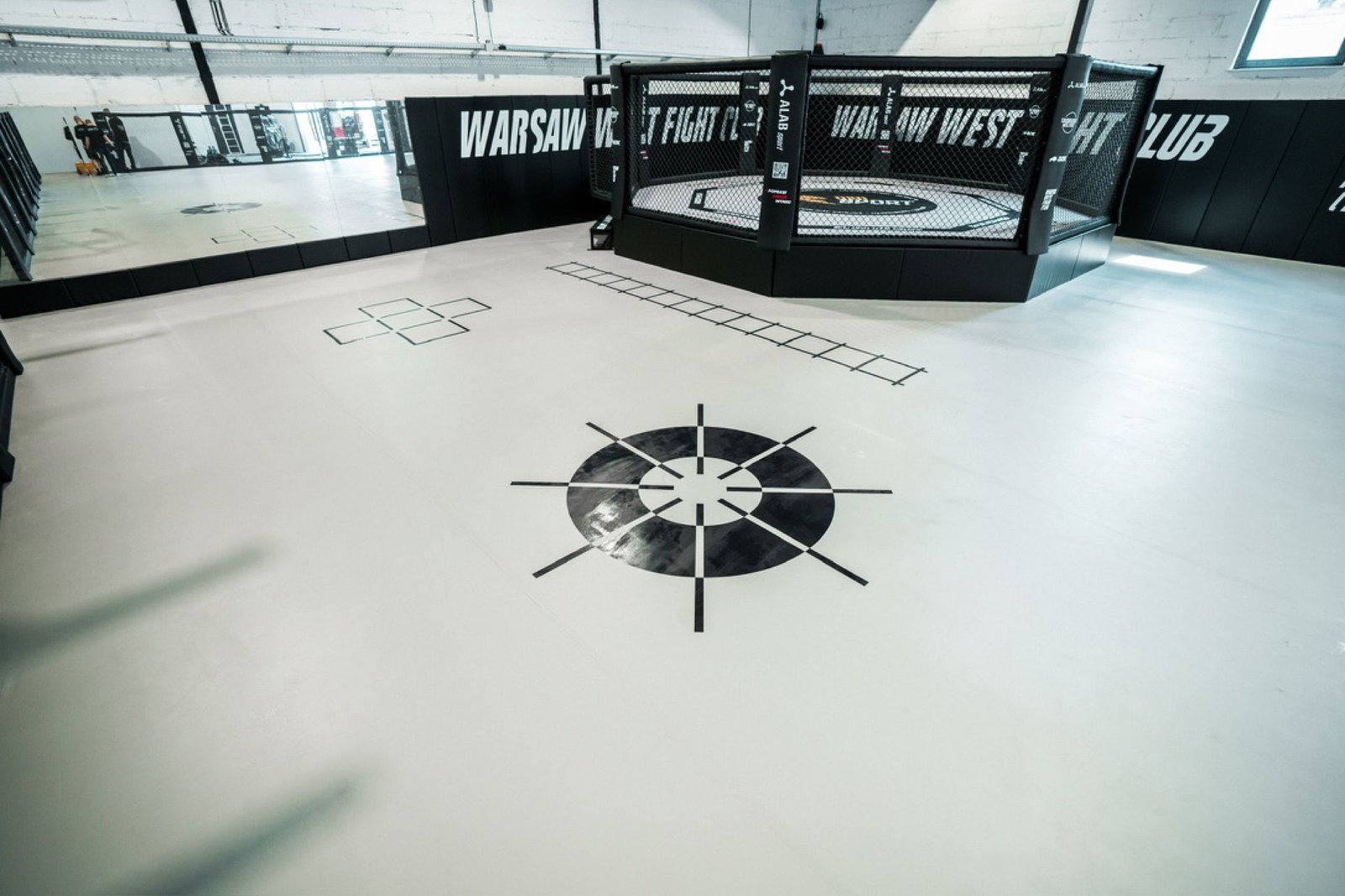 Koło footwork i drabinka nadrukowane na jasnej macie przed klatką MMA na podeście