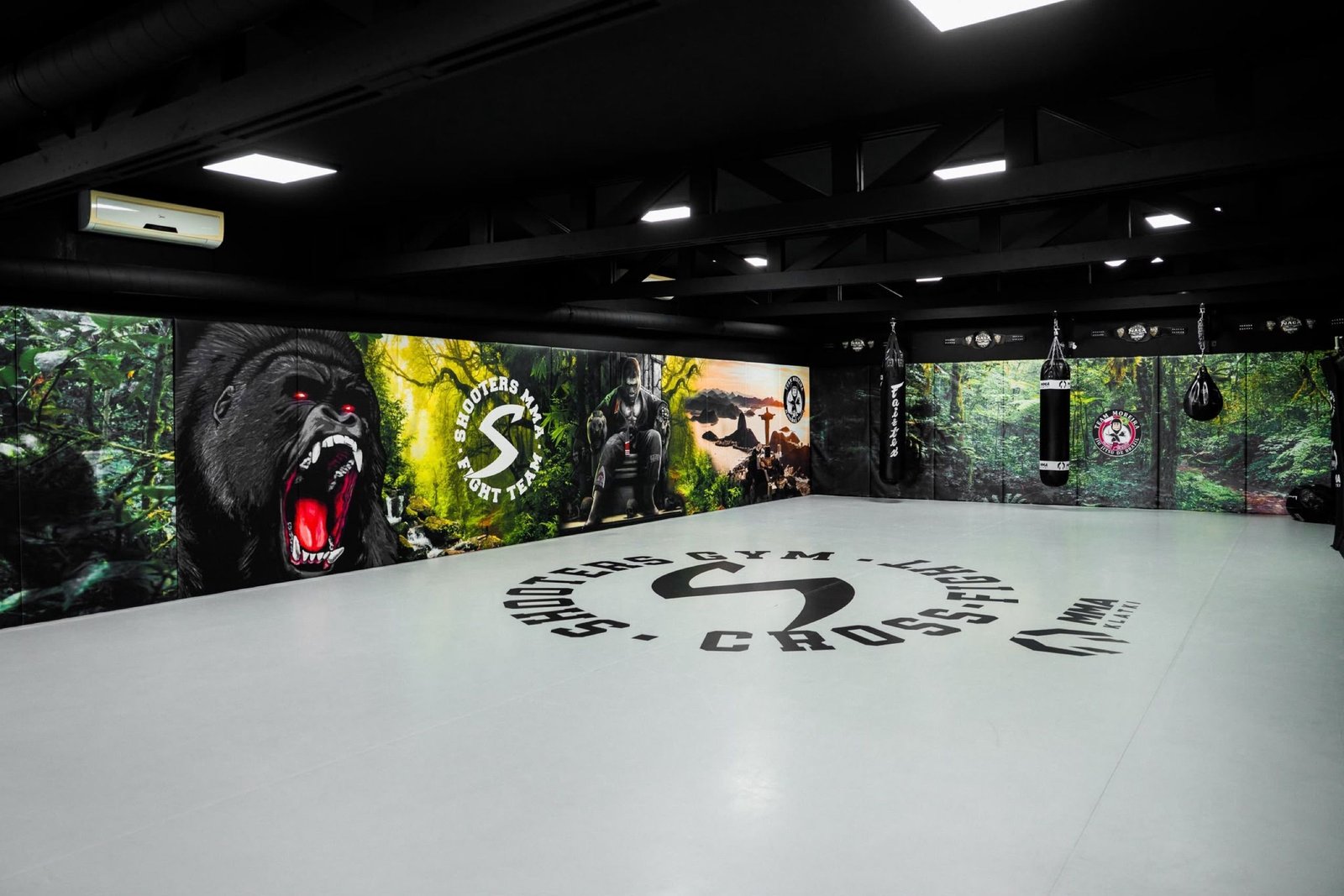 Sala Shooters Gym z centralnym logo na jasnej macie, muralami dżungli na matach ściennych i strefą worków