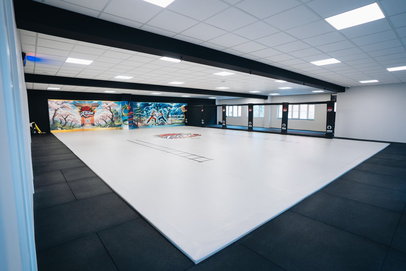 Szerokie ujęcie sali Róża Fight Team z narożnika. Jasna mata treningowa z logo klubu łączy się z matami ściennymi i czarnym systemem Cage Wall.