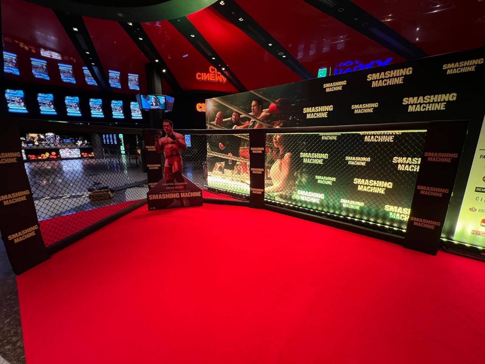 Poziome ujęcie ukończonej strefy fotograficznej w foyer kina. Szeroki czerwony dywan prowadzi do konstrukcji złożonej z klatki MMA z czarnej siatki po lewej stronie i podświetlonej ścianki sponsorskiej po prawej, pokrytej gęsto powtórzonym logotypem „SMASHING MACHINE" oraz dużymi kadrami z filmu (scena walki w ringu, publiczność). Pośrodku stoi kartonowy standee zawodnika MMA z tytułem filmu i nazwiskami aktorów. Sufit ma czerwono-czarną, pasiastą formę, świecą neony „CINEMA CITY" i „IMAX", a na ścianach widoczne są podświetlane plakaty i ekrany repertuarowe. Po prawej stronie kadru widać fragment roll-upu z logotypami partnerów.