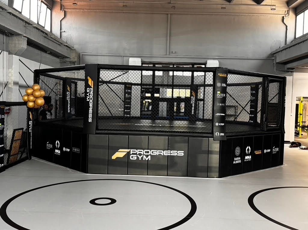 Ośmiokątna klatka MMA na podeście z brandingiem klubu w sali treningowej, szara mata z kołami zapaśniczymi