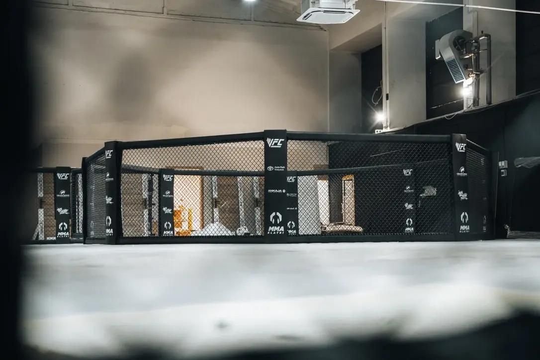 Klatka MMA na poziomie posadzki w przestronnej hali, czarne obicia słupów z logotypami sponsorów