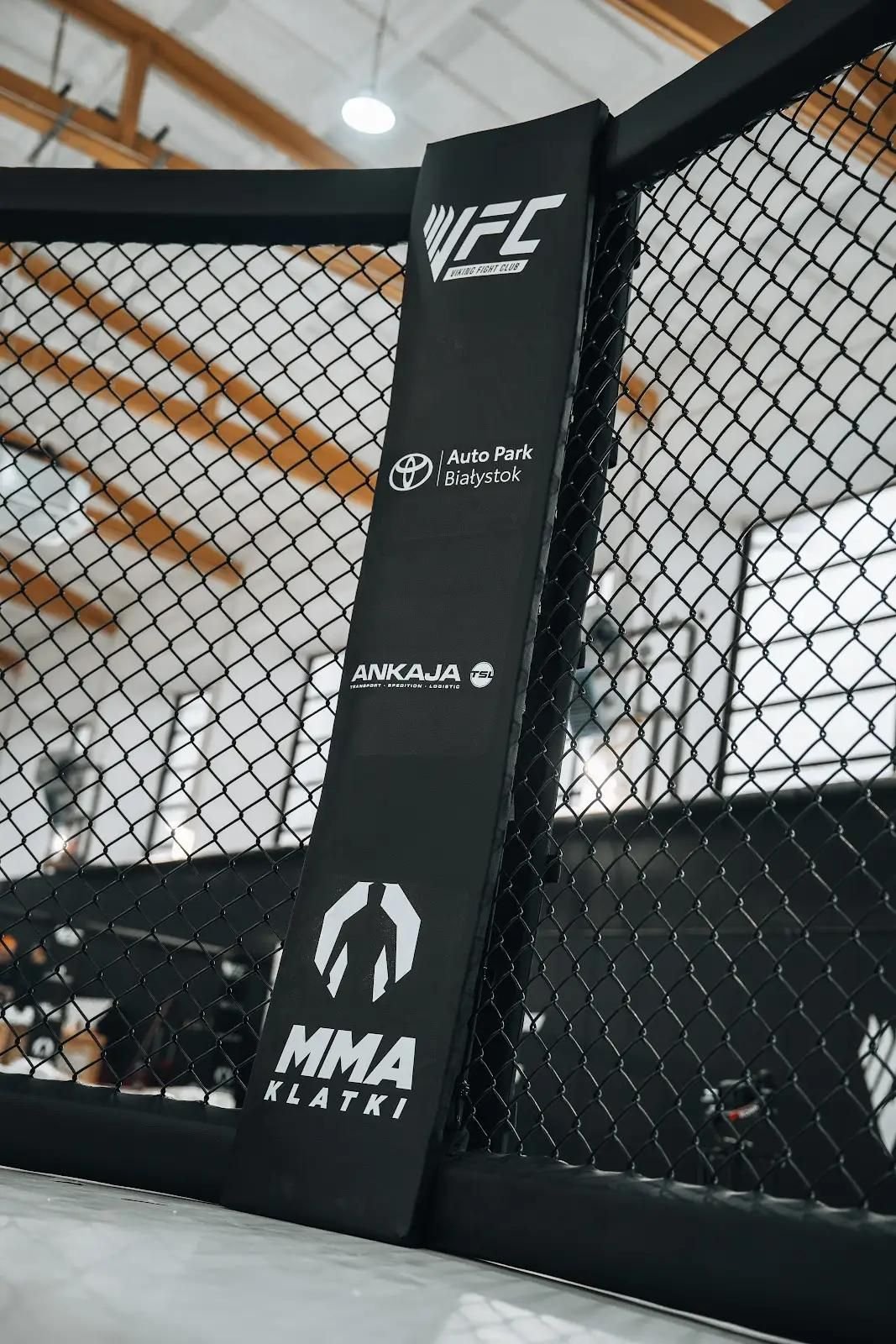 Biała mata klatki MMA z nadrukiem logo, zbliżenie przez siatkę panelu z czarnymi słupami w tle