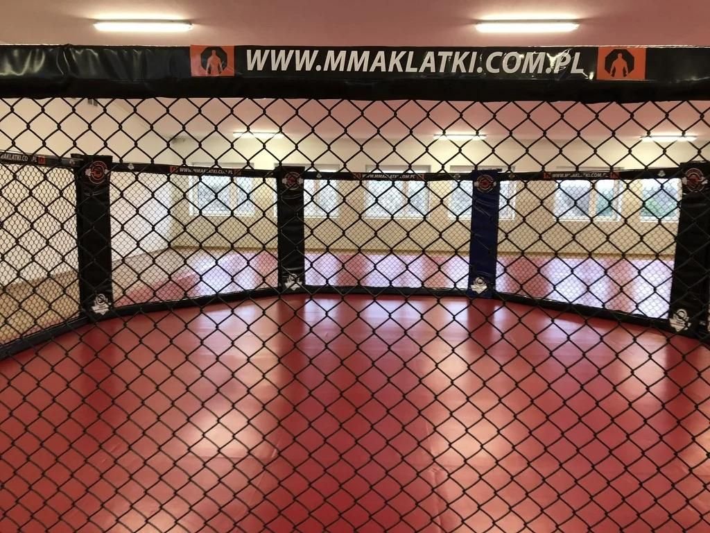 Wnętrze klatki MMA CLASSIC widziane przez siatkę: czarne obicia słupów, górna banda z nadrukiem, czerwona mata i okna sali