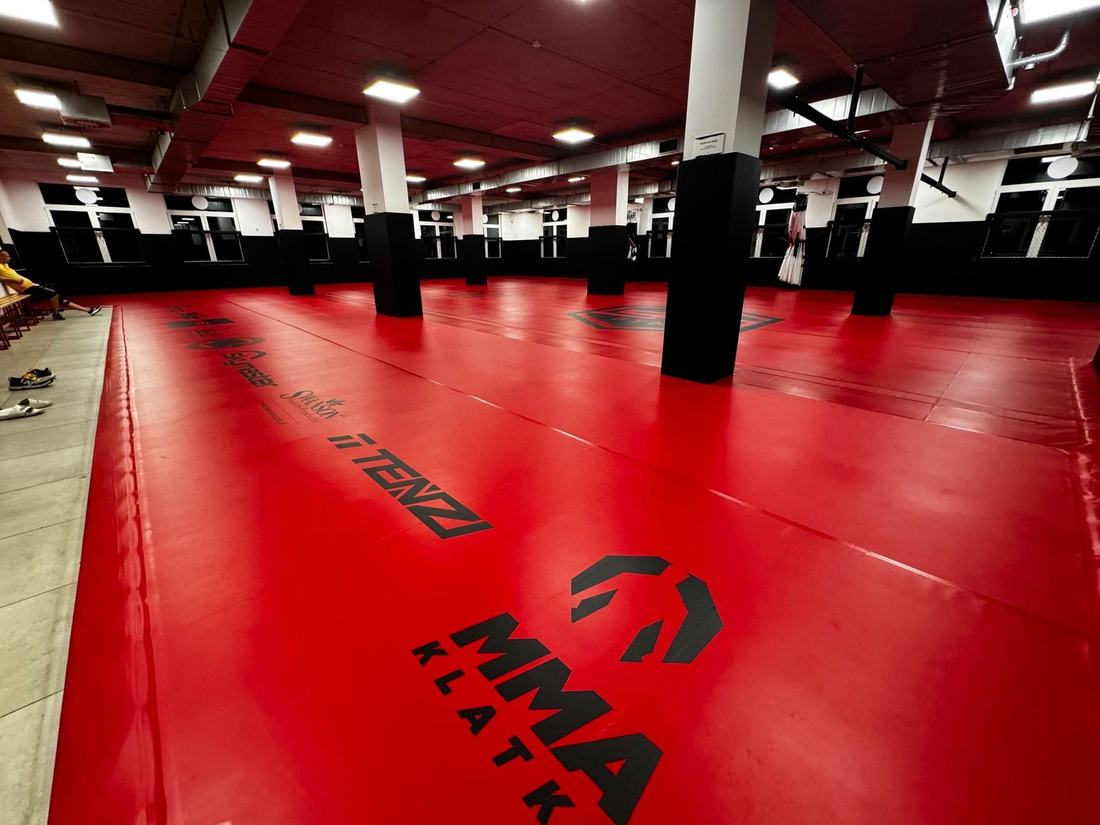 Czerwona mata z nadrukami sponsorów między czarnymi filarami sali Open Space BT Gym