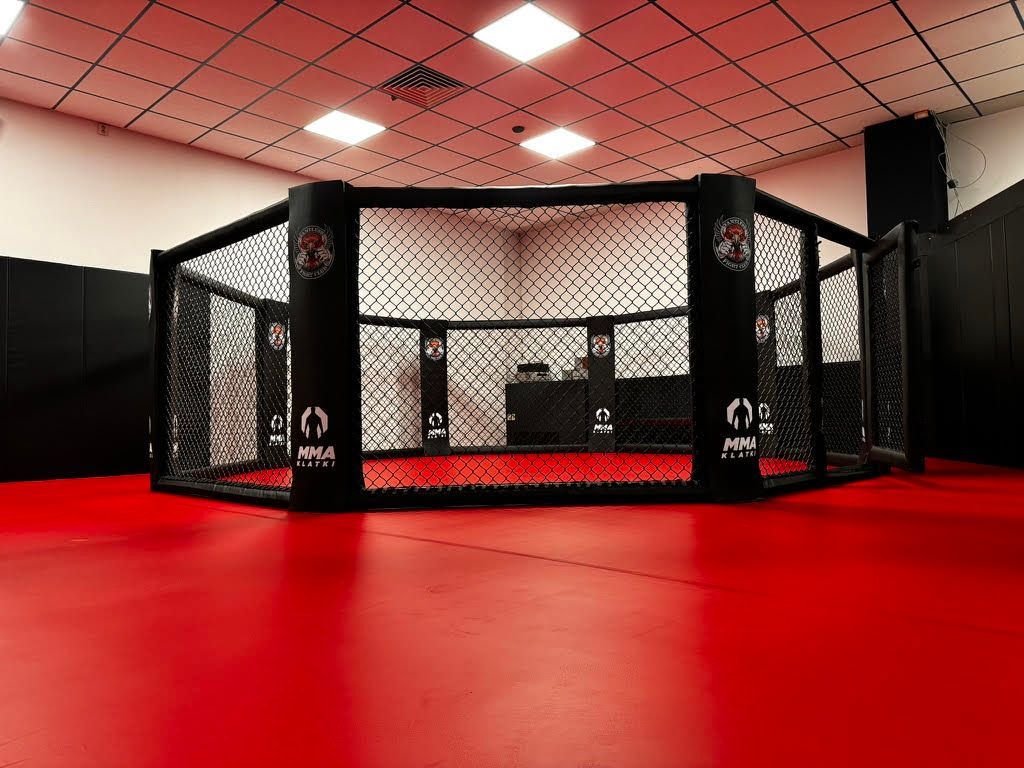 Ośmiokątna klatka MMA na czerwonej macie w sali klubowej, czarne obicia słupów i panele z siatką