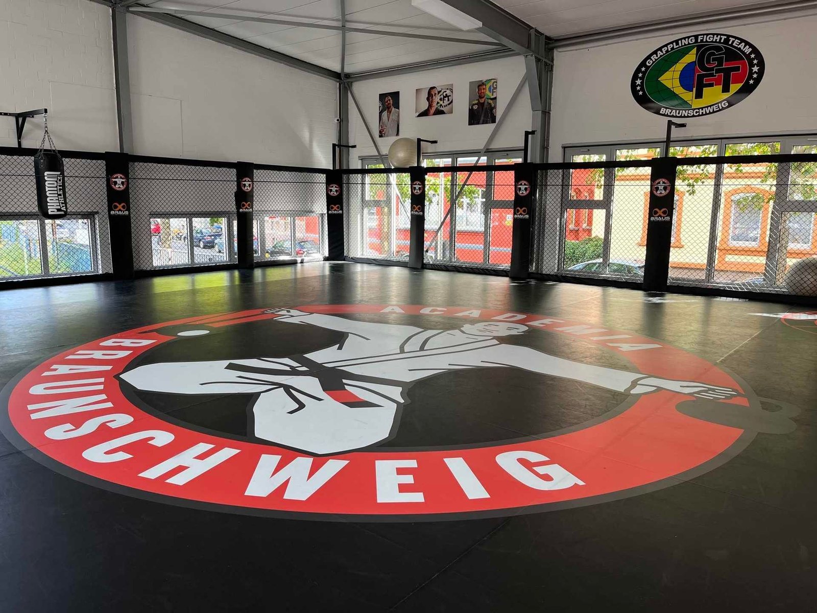 Panoramiczne ujęcie jasnej hali treningowej o wysokim, białym stropie ze stalową konstrukcją. Podłogę pokrywa ciemna mata treningowa z dużym okrągłym nadrukiem „ACADEMIA BRAUNSCHWEIG" w kolorze czerwono-białym. Powierzchnia treningowa otoczona jest ogrodzeniem z czarnej siatki, którego słupy osłonięto czarnymi ochraniaczami z logo klubu i marki BRAUS. Po lewej stronie na łańcuchu wisi czarny worek bokserski Phantom Athletics. Na białej ścianie w tle znajdują się trzy plakaty ze zdjęciami zawodników oraz duży owalny szyld „GRAPPLING FIGHT TEAM BRAUNSCHWEIG". Za ogrodzeniem, przez przeszklenia, widać parking z samochodami i drzewa.