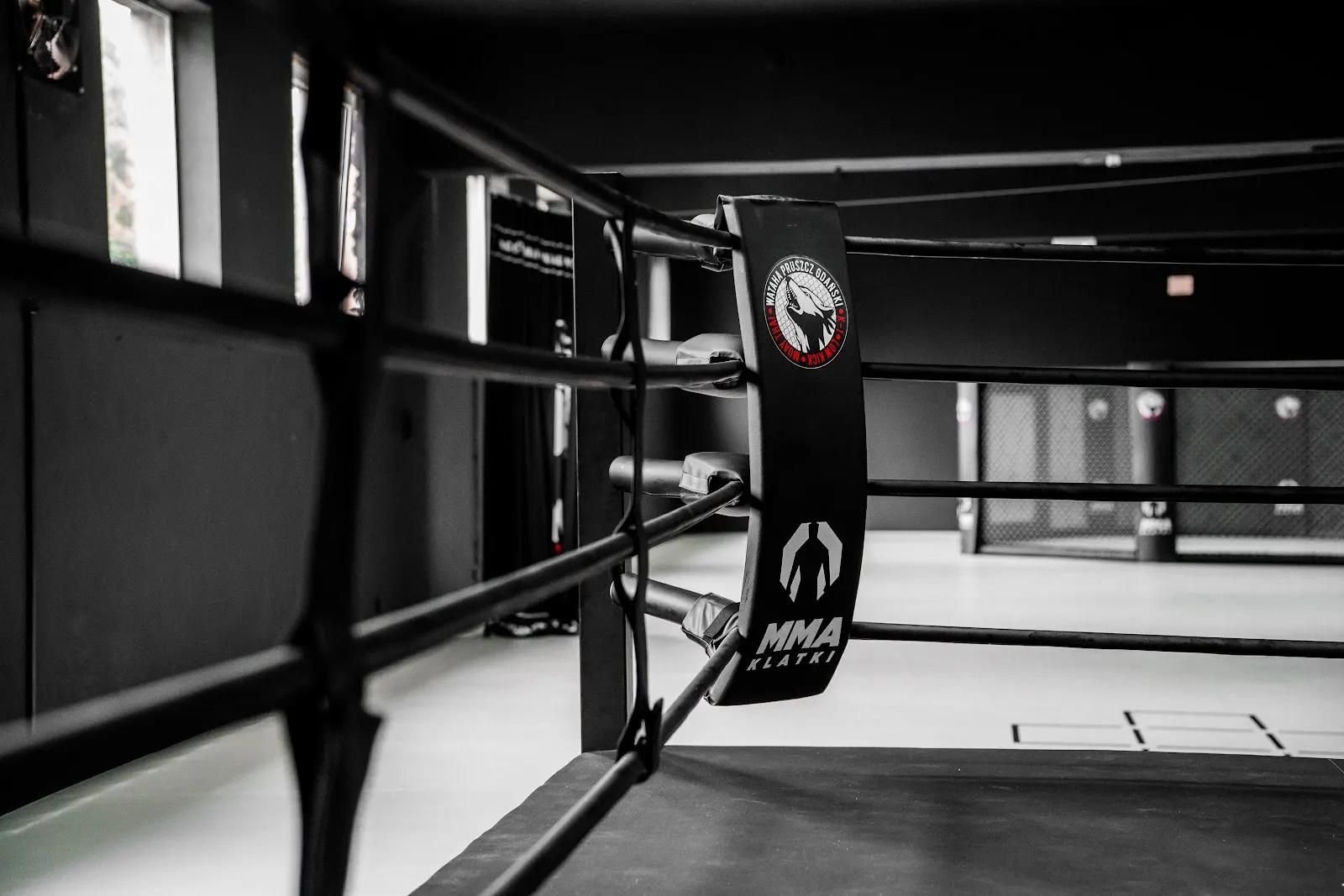 Ring bokserski z boku: czarna poduszka narożnika z logo klubu i cztery liny w ciemnej hali