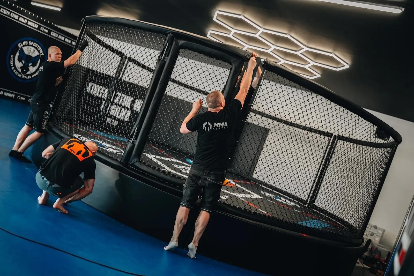 Montaż ściany z siatki przez monterów, niebieska podłoga, heksagonalne oświetlenie, wyposażenie sali MMA w trakcie realizacji