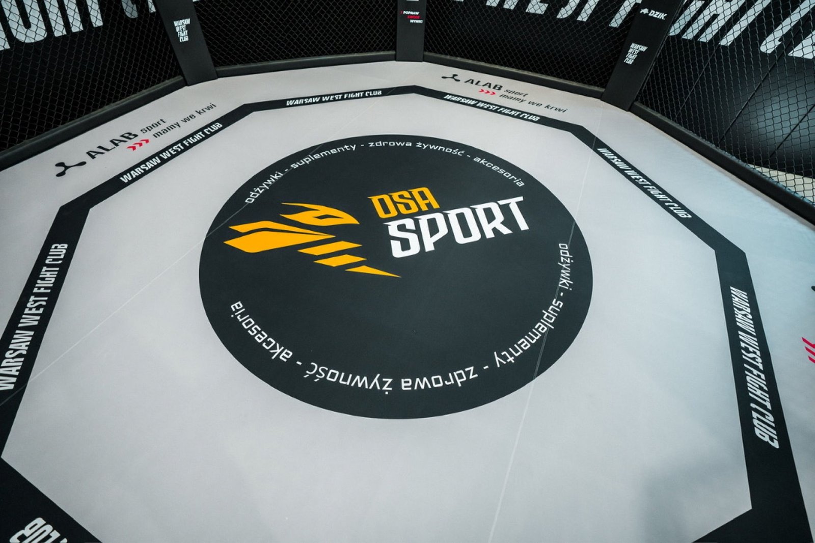 Wnętrze klatki Warsaw West Fight Club z matą z logo sponsora i obszyciami z nazwą klubu