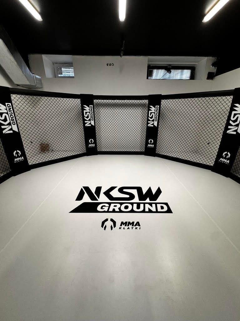 Wnętrze klatki MMA z białą matą i logo klubu, czarne słupy z siatką w niskiej sali klubowej