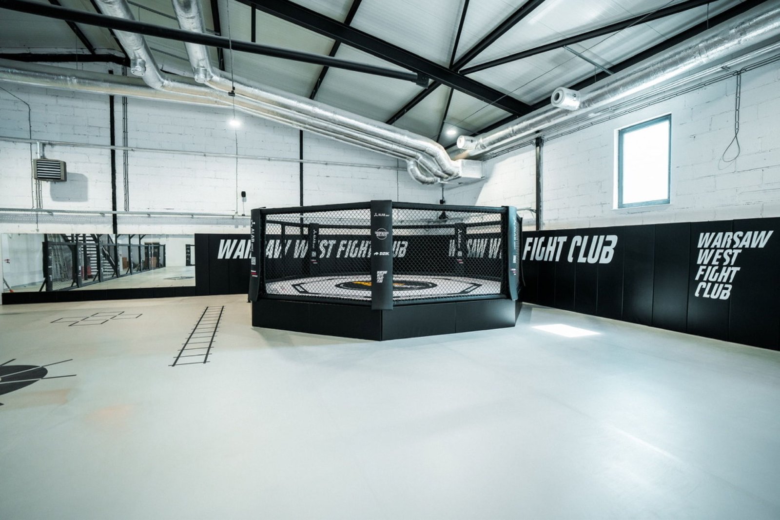 Klatka MMA o średnicy 7 m na podeście z napisami Warsaw West Fight Club na matach ściennych