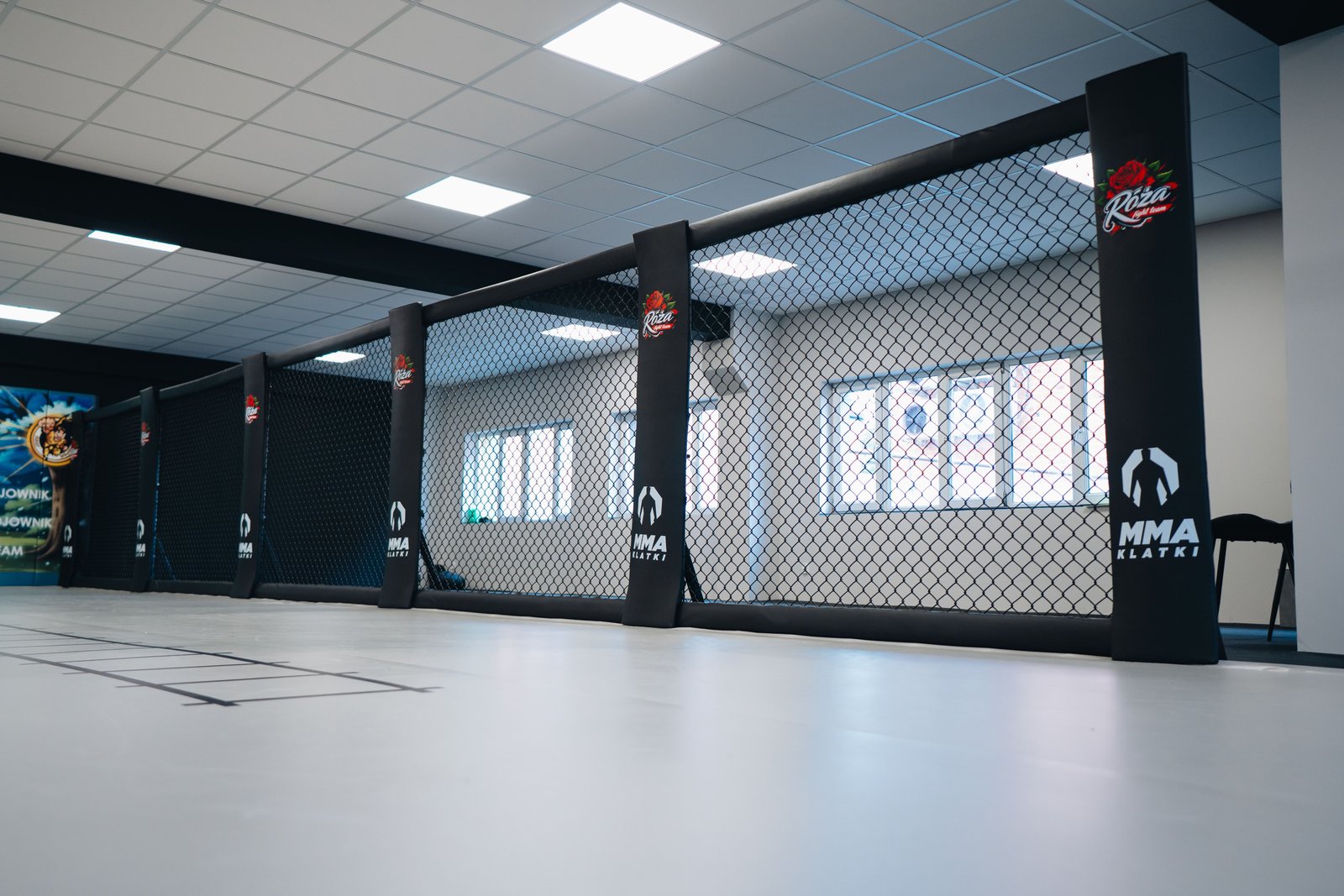 System Cage Wall w sali Róża Fight Team. Czarne panele z siatką mają osłonięte słupy z logo klubu i MMA Klatki.