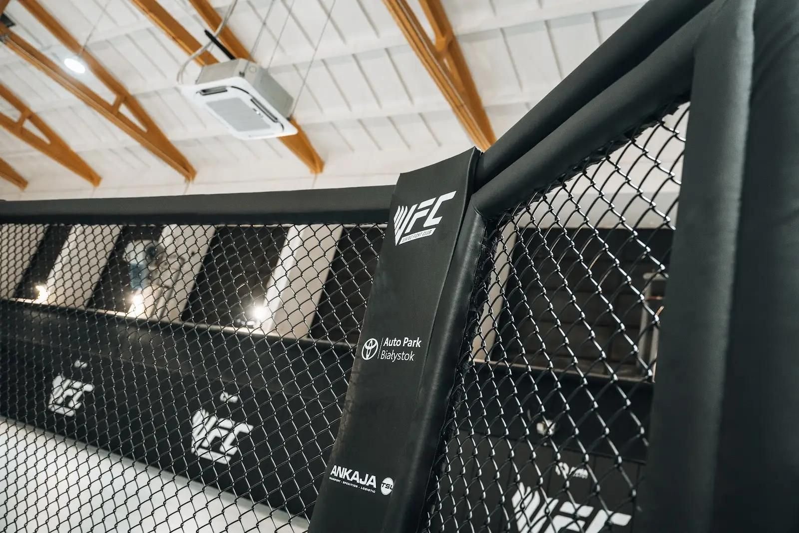 Zbliżenie obitego słupa narożnego klatki MMA i górnej bandy, siatka i drewniane dźwigary hali