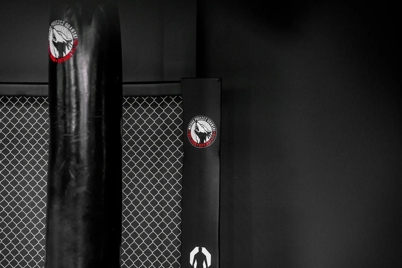 Czarny worek treningowy z logo klubu obok osłony słupka Cage Wall, siatka na tle czarnej ściany