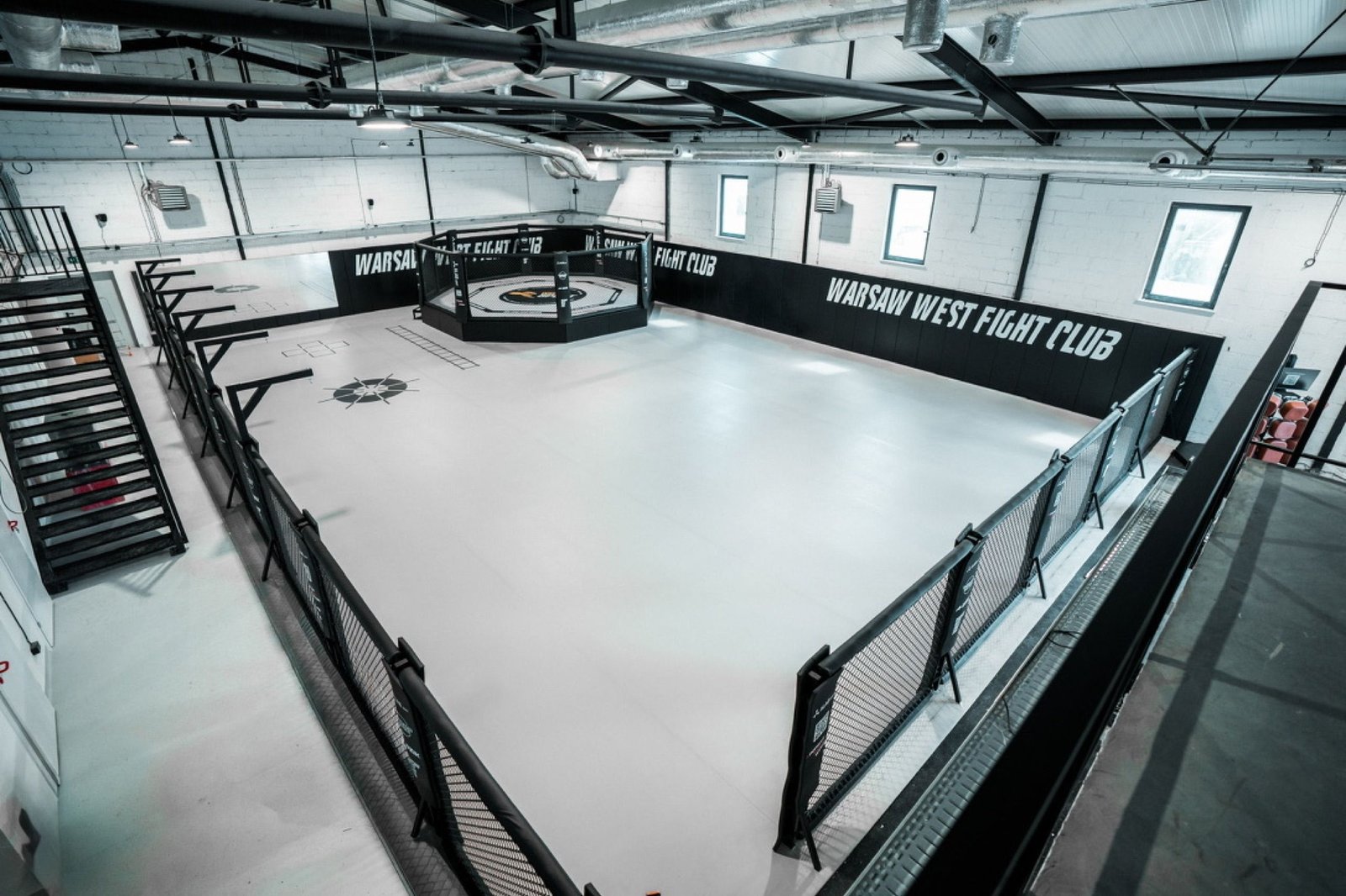 Widok z antresoli na główną strefę Warsaw West Fight Club z klatką MMA na podeście i jasną matą