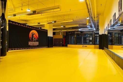 Żółta mata podłogowa w sali klubu Komodo z banerem klubowym, klatką MMA i workami bokserskimi