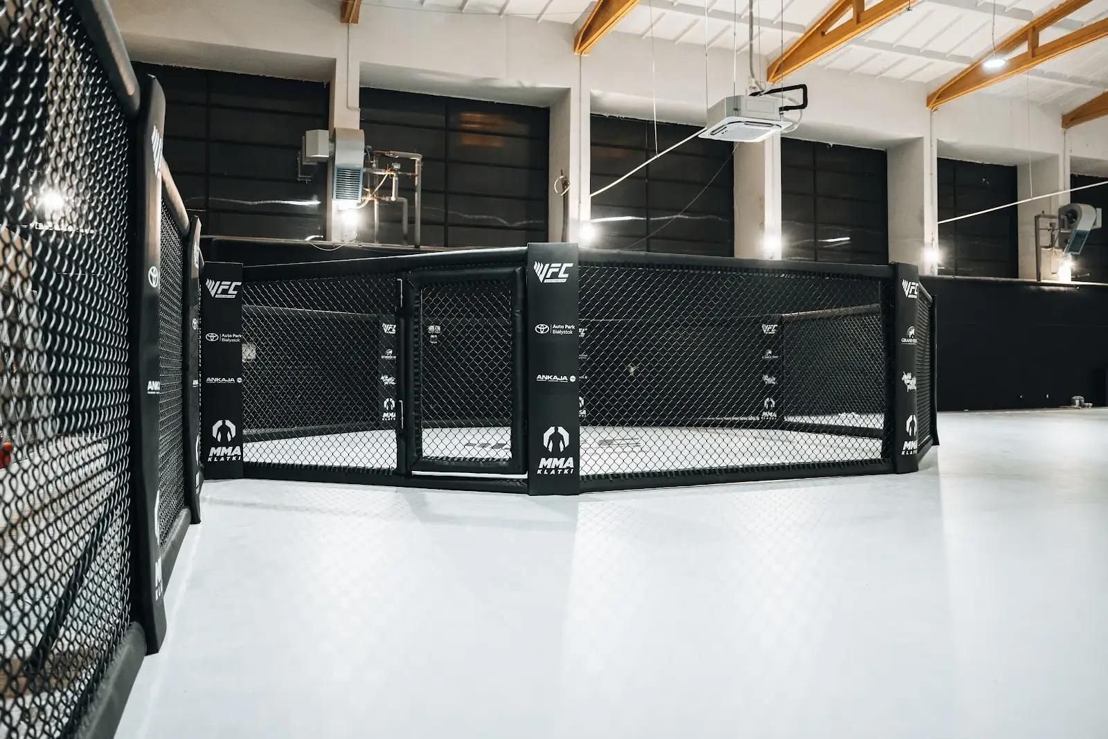 Klatka MMA z drzwiami wejściowymi na jasnej macie w hali sportowej, panele z siatką i obite słupy