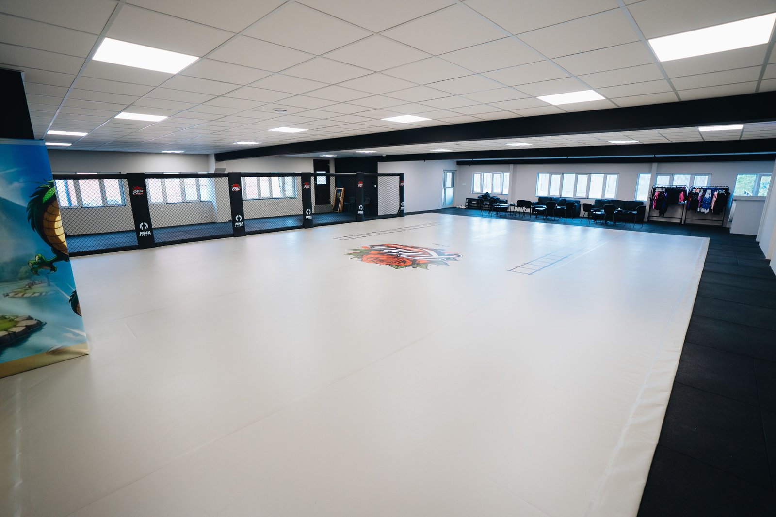 Widok sali Róża Fight Team od strony mat ściennych. Na pierwszym planie jest jasna mata z logo, a w tle system Cage Wall i strefa wypoczynkowa.