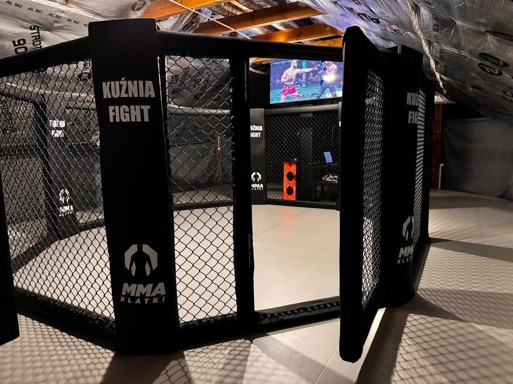 Klatka MMA z otwartymi drzwiami w sali treningowej na poddaszu, szare maty i czarne obicia słupów