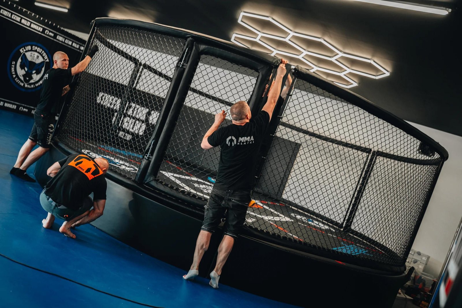 Montaż klatki MMA na podeście w Strefie Sztuk Walki Mogilno: ekipa MMA Klatki naciąga nową siatkę, niebieska mata, czarne maty ścienne z logo klubu