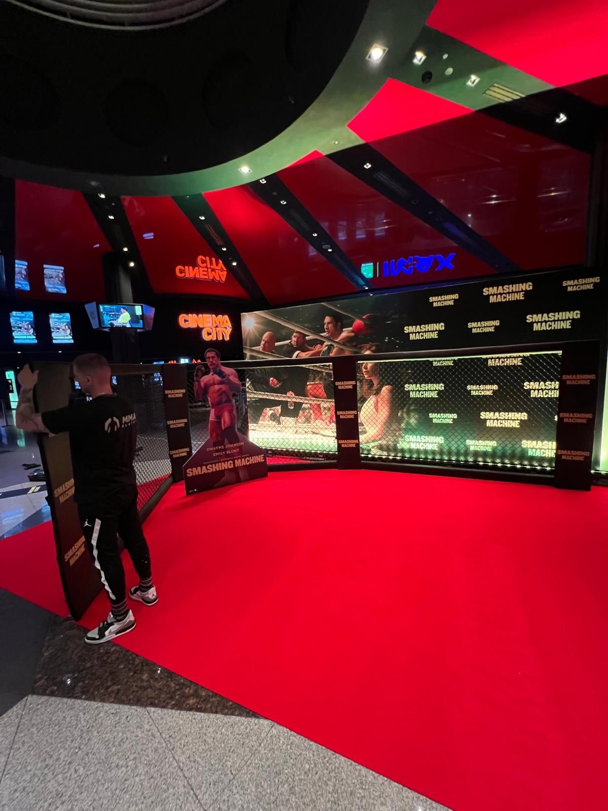 Szerokie ujęcie foyer Cinema City z charakterystycznym czerwono-czarnym sufitem i neonami „CINEMA CITY" oraz „IMAX". Na czerwonym dywanie ustawiona jest strefa fotograficzna: klatka MMA z czarnej siatki połączona z podświetloną ścianką sponsorską pokrytą powtarzającymi się napisami „SMASHING MACHINE" i kadrami z filmu. Pośrodku stoi kartonowy standee (figura reklamowa) przedstawiający zawodnika MMA w czerwonych spodenkach, z podpisem „DWAYNE JOHNSON, EMILY BLUNT — SMASHING MACHINE". Po lewej stronie mężczyzna w czarnej bluzie z logo MMA Klatki przenosi czarny element ochraniacza. W tle widoczne są kasy i ekrany z repertuarem kina.