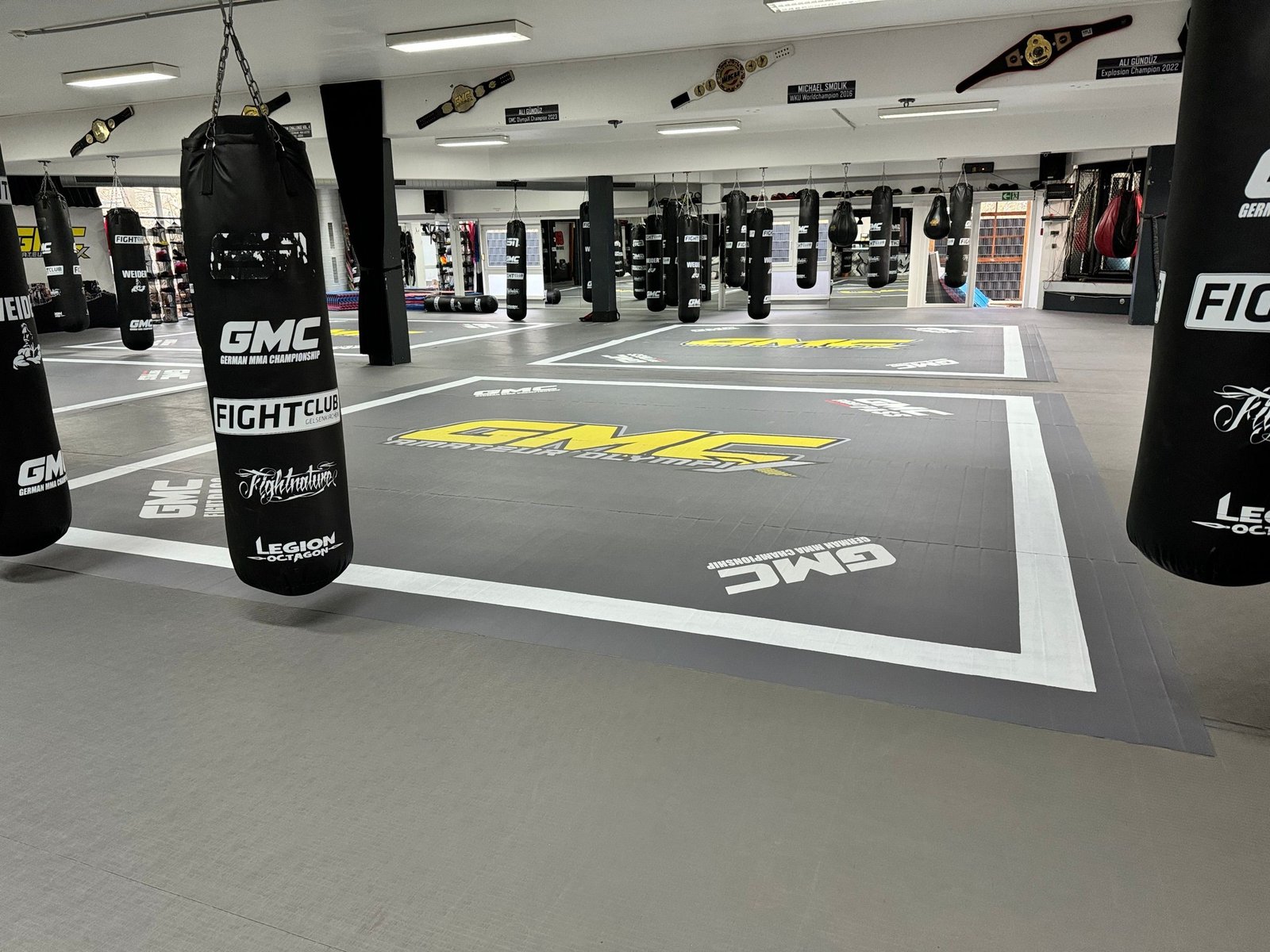 Sala GMC z szarymi matami z nadrukami klubu, strefą worków bokserskich i klatką MMA w tle