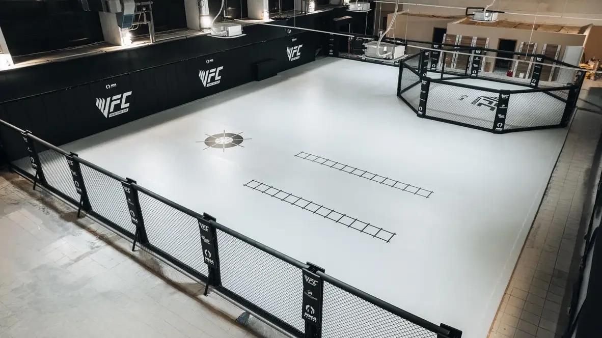 Widok z góry na wyposażenie sali MMA: biała mata, czarne materace ścienne, ściana z siatki i klatka